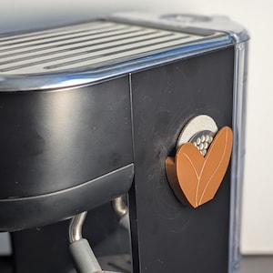 Könnte beinhalten: Schwarz-silberne Espressomaschine mit Dampflanze und Tropfschale aus Edelstahl. Die Maschine hat eine silberne Oberseite und einen dekorativen braun-weißen blattförmigen Akzent. Der Markenname "Sage" ist sichtbar.