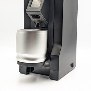 Op de afbeelding: Een zwarte koffiemolen met een zilverkleurige metalen doseerbeker aan de voorzijde. Het apparaat is voorzien van een grijze balg aan de bovenzijde, een vierkant digitaal display en een kleine instelknop op de bovenste behuizing.