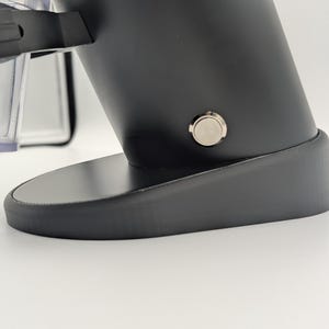 May include: A black electric coffee grinder with a silver button and a clear plastic container. The grinder has a sleek, modern design and sits on a rounded base. The image is taken on a white surface.
