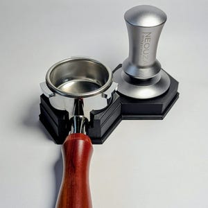 Puede incluir: Un portafiltro de acero inoxidable y un tamper de espresso plateado con el texto "NEOUZA espresso" se muestran. El portafiltro tiene un mango de madera. Ambos artículos están sobre una base hexagonal negra.