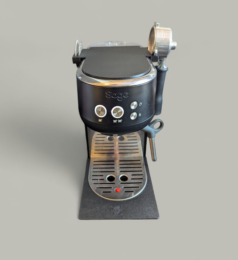 May include: Black and silver espresso machine with the brand name "Sage". The machine has a stainless steel drip tray and a portafilter. It features several buttons and a dial for operation.