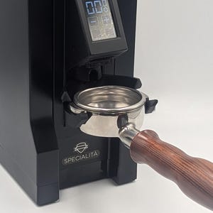 Könnte beinhalten: Eine schwarze Espressomühle mit einem digitalen Display, das 0.0 anzeigt, und einem Siebträger mit poliertem Metallkorb sowie einem dunkelbraunen Holzgriff. Auf der Vorderseite der Maschine steht der Text SPECIALITA mit einem Schildsymbol.