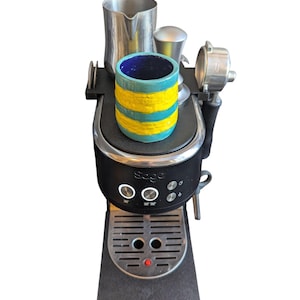 May include: A black espresso machine with a stainless steel portafilter, milk frothing jug, and a small cup. The cup is cylindrical, with a blue interior and a turquoise and yellow striped exterior. The machine has the brand name "Sage" on it.