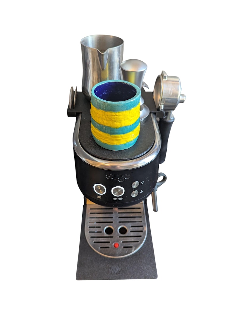 May include: A black espresso machine with a stainless steel portafilter, milk frothing jug, and a small cup. The cup is cylindrical, with a blue interior and a turquoise and yellow striped exterior. The machine has the brand name "Sage" on it.