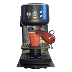 Puede incluir: Máquina de espresso negra con la marca "Sage" en la parte superior. La máquina tiene botones plateados y un mango de madera. Una taza naranja se encuentra en la bandeja de la máquina y debajo hay una báscula digital.