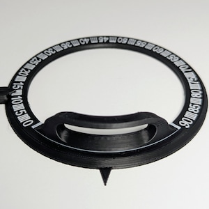 Puede incluir: Una herramienta circular negra con marcas blancas y un asa. La herramienta tiene una punta y medidas de 5 a 90, probablemente para medir ángulos o distancias. La herramienta está hecha de un material negro mate.