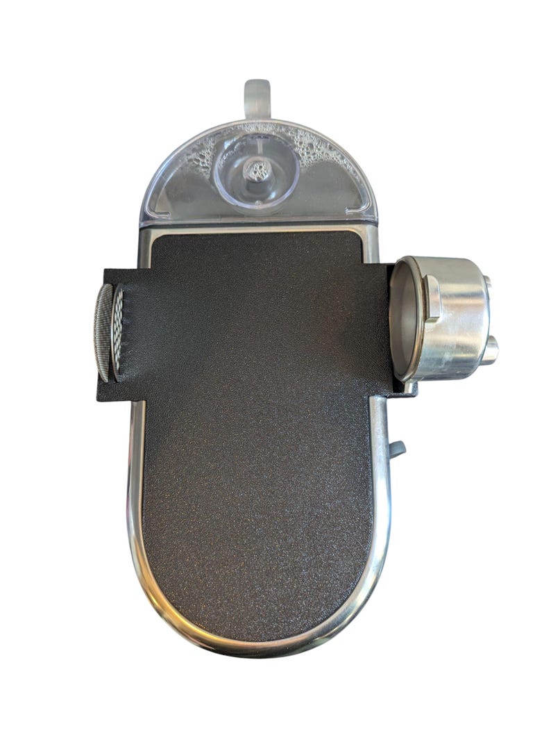 May include: A black and silver espresso machine, viewed from above. It features a clear water reservoir, a textured black surface, and a silver metal portafilter. The design is modern and compact, suitable for making coffee at home or in the office.