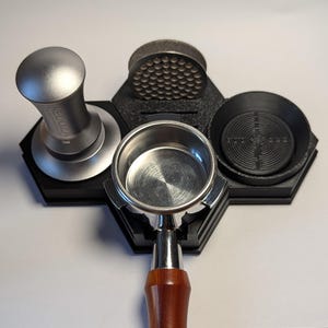 Peut inclure: Un organisateur de poste de café noir avec un tamper argenté, un outil de distribution, un porte-filtre et un bac à marc. Le manche du porte-filtre est en bois. L'organisateur est sur une surface blanche.