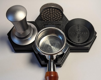 May include: A black coffee station organiser with a silver tamper, a distribution tool, a portafilter holder, and a knock box. The portafilter handle is made of wood. The organiser is on a white surface.