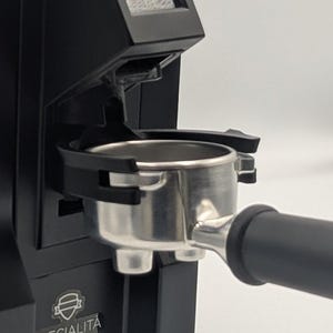 Puede incluir: Un molinillo de café negro con un portafiltros metálico insertado en el soporte frontal. La máquina tiene una pequeña pantalla digital en la parte superior y la palabra SPECIALITA impresa en blanco sobre un pequeño logo en la parte inferior.