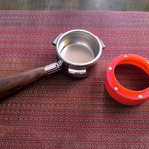 May include: Espresso machine portafilter with a wooden handle and a stainless steel filter basket. An orange tamper ring with white dots is next to the portafilter. The items are on a textured, reddish-brown surface.