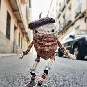 Könnte beinhalten: Eine gehäkelte Eichelpuppe mit bunten, gestreiften Socken und Rollschuhen. Die Puppe steht auf einer grauen Asphaltstraße.