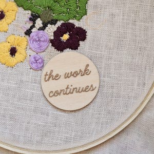May include: A close-up of a wooden embroidery hoop with a beige fabric background. The fabric is embroidered with purple and yellow flowers. A wooden circle with the text "the work continues" is attached to the fabric.