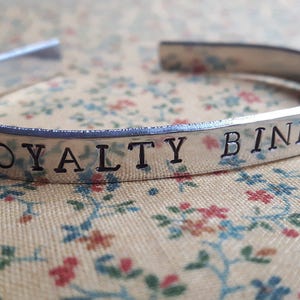 May include: Silver bracelet with the inscription "Loyalty Binds" on a floral patterned background.