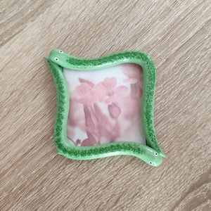 May include: A square picture frame with a green snake design. The frame is light green with darker green detailing and two small, white eyes on each snake head. The frame holds a picture with pink flowers.