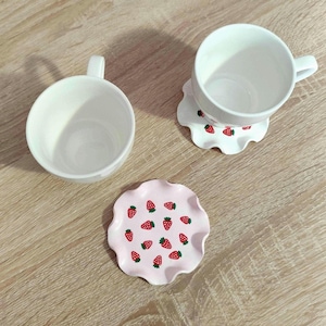 Op de afbeelding: Twee witte keramische mokken en twee bijpassende onderzetters. Eén onderzetter zit onder een mok en de andere naast de tweede mok. De onderzetters en de randen van de mokken hebben een aardbeienpatroon.