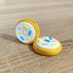 Op de afbeelding: Twee gele, ronde, decoratieve objecten die op koekjes lijken. Elk heeft een witte glazuurlaag met kleurrijke hagelslag in roze, blauw en geel. De objecten staan op een houten oppervlak.