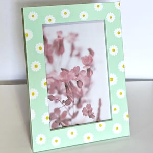 Può includere: Una cornice per foto verde chiaro con un motivo di margherite bianche attorno al bordo. La cornice contiene una foto di fiori rosa.
