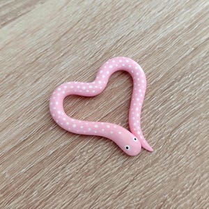 May include: A pink snake-shaped figurine with white polka dots, forming a heart shape. The snake has small black eyes and a slightly raised tail. The figurine is set against a light brown wooden surface.