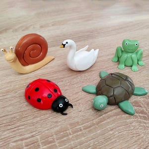 Può includere: Una collezione di cinque figurine di animali colorate: una lumaca, un cigno, una rana, una coccinella e una tartaruga. La lumaca ha un guscio marrone e un corpo beige. Il cigno è bianco con un becco giallo. La rana è verde. La coccinella è rossa con pois neri. La tartaruga è marrone e verde.
