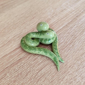 Op de afbeelding: Handgemaakte groene slangenfiguur met een gespikkeld patroon. De slang is opgerold, met een kleinere slangenkop erbovenop. Het beeldje is gemaakt van glanzend materiaal en staat op een houten achtergrond.