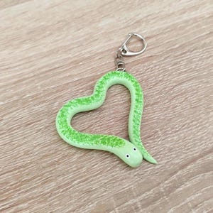 Può includere: Un portachiavi a forma di serpente verde chiaro piegato a cuore. Il serpente presenta una texture con un motivo maculato in una tonalità di verde più scura, due piccoli occhi neri sulla testa ed è fissato a un anello in metallo argentato.
