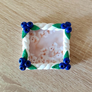 May include: A square picture frame with a white braided border. The corners are decorated with clusters of blue blueberries and green leaves. The frame's interior displays a soft, floral background.
