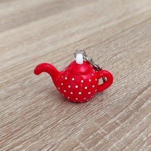Könnte beinhalten: Ein roter Miniatur-Teekannen-Schlüsselanhänger mit weißem Punktemuster auf dem abgerundeten Körper. Die Teekanne hat einen geschwungenen Ausguss, einen kleinen Henkel und einen weißen Knauf auf dem Deckel, mit einem silbernen Metallring oben.