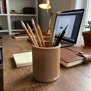 Pode incluir: Um porta-lápis cilíndrico com textura canelada vertical, feito de um material castanho claro. O suporte contém vários lápis de madeira com borrachas cor-de-rosa, uma caneta de ponta fina clara e uma lapiseira preta.