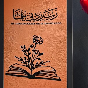 May include: Framed artwork with a textured orange background. The artwork features Arabic calligraphy and the English text "MY LORD INCREASE ME IN KNOWLEDGE." A black floral design is over an open book. The frame is black.