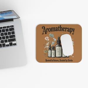 May include: A brown mouse pad with the text "Aromatherapy" in black lettering. The text "Rooted in Nature, Healed by Herbs" is below the main text. An illustration of five bottles of essential oils and flowers is in the center of the mouse pad.