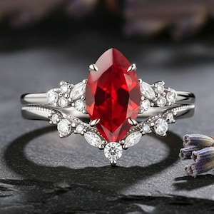 May include: A two-piece ring set featuring a marquise-cut, deep red gemstone as the focal point. The main ring is flanked by smaller, clear gemstones. The band is silver-toned, and the set is displayed on a dark surface.