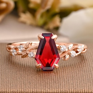 Puede incluir: Anillo de oro rosa con una gema roja oscura en forma de ataúd. La banda tiene un diseño floral con pequeñas piedras claras. El anillo está sobre una superficie marrón texturizada.