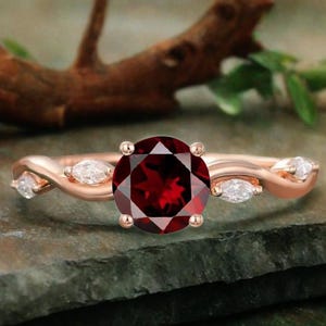 Puede incluir: Anillo de oro rosa con una gran gema redonda de color rojo intenso. La banda tiene un diseño retorcido con pequeñas piedras blancas en forma de marquesa. El anillo se presenta sobre un fondo natural, resaltando su elegante diseño.