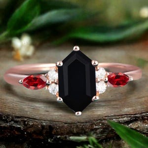 May include: Rose gold ring featuring a large, black, hexagonal center stone. Accented with small white stones and two red, marquise-cut stones. The ring is set against a natural wood background with green foliage.