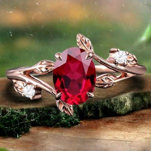 May include: Rose gold ring featuring an oval, deep red gemstone. The ring's band is designed with delicate leaf and vine details, and accented with two small, clear gemstones. The ring is set against a blurred natural background.