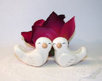 May include: Two white ceramic doves with closed eyes and smiling faces are perched on a white doily. A single burgundy rose is behind the doves.
