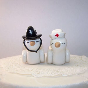 Pode incluir: Um par de enfeites de bolo de casamento extravagantes. Uma figura usa uma cartola preta e a outra um gorro de enfermeira com uma cruz vermelha. Ambas são brancas com bicos castanhos e expressões sorridentes. Estão sobre uma toalha de renda branca.