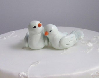 May include: Two small, light blue bird figurines with orange beaks sit atop a white cake. The birds are facing each other, suggesting a wedding cake topper. The cake has a white, smooth surface with delicate floral patterns.