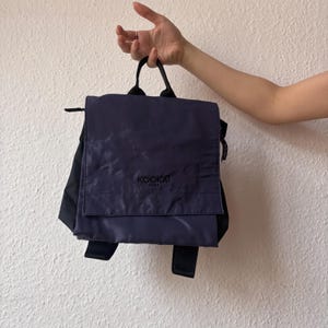 Può includere: Uno zaino blu scuro con spalline nere e un logo Kookai Paris. Lo zaino ha una chiusura a lembo e una tasca con cerniera sul davanti.