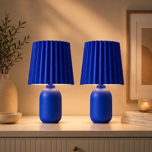 May include: Two matching blue table lamps with pleated shades. The lamps have a rounded base and a vibrant blue color. The lamps are placed on a white surface, with a neutral background.