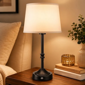 May include: A table lamp featuring a matte black metal base with a slender, tiered central column and a circular foundation. The lamp is topped with a tapered, off-white fabric drum shade that emits a soft, warm glow from the internal light source.