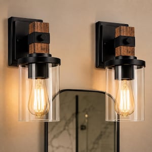 May include: Two wall-mounted light fixtures featuring a rectangular black metal backplate, a vertical brown wood-textured block, and a cylindrical clear glass shade. Each fixture contains a single Edison-style light bulb emitting a warm glow.