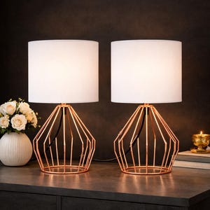 May include: Two modern table lamps with white cylindrical shades and geometric copper-colored metal bases. The lamps are placed on a dark surface, with a vase of flowers and a candle in the background. The lamps are connected by a black cord.