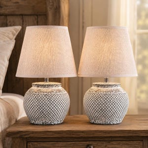 May include: A pair of table lamps featuring round, textured ceramic bases with a white and dark gray cross-hatch pattern and a distressed finish. Each lamp is topped with a tapered, light beige fabric drum shade that emits a warm glow.