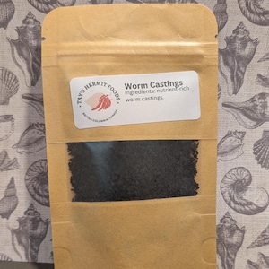 May include: A brown paper bag with a clear window showing black worm castings. The bag has a label that reads "Tay's Hermit Foods" and "Worm Castings. Ingredients: nutrient-rich worm castings."