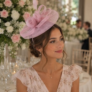 May include: A dusty pink fascinator hat featuring a wide, sheer, circular brim with structured loops and a single upright feather detail. The headpiece is secured on a thin headband, showcasing a delicate, semi-transparent fabric construction.
