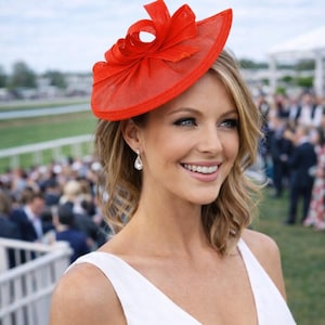 Peut inclure: Une femme porte un chapeau rouge vif orné d'un nœud. Elle porte également une robe blanche et des boucles d'oreilles en forme de goutte. L'arrière-plan montre une foule de personnes et une zone herbeuse, suggérant un événement en plein air.