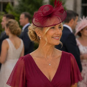 Op de afbeelding: Een diepbordeauxrode fascinator met een brede, ronde sinamay basis, gestructureerde lussen en rechtopstaande veren. Het hoofddeksel wordt gedragen bij een bijpassende bordeauxrode jurk met V-hals en transparante, wapperende korte mouwen.