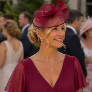 Puede incluir: Un tocado fascinator de color burdeos intenso con tela de malla transparente en capas y detalles de plumas erguidas, combinado con un vestido burdeos a juego con escote en V y mangas transparentes, y un delicado collar de cadena plateada.